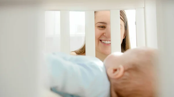 Портрет счастливой смеющейся женщины, смотрящей и держащей своего ребенка в кроватке. Концепция воспитания, семейного счастья и развития ребенка — стоковое фото