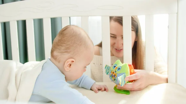 Moeder zit aan de wieg en geeft speelgoed aan haar zoontje. Begrip ouderschap, gezinsgeluk en ontwikkeling van baby 's — Stockfoto