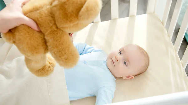 Madre dando peluche oso de peluche a su pequeño bebé acostado en la cuna. Concepto de paternidad, felicidad familiar y desarrollo del bebé — Foto de Stock