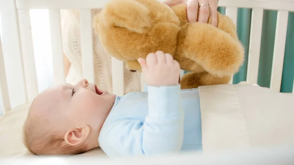 Feliz niño sonriente palideciendo y sosteniendo oso de peluche mientras está acostado en una cuna de madera. Concepto de paternidad, felicidad familiar y desarrollo del bebé — Foto de Stock