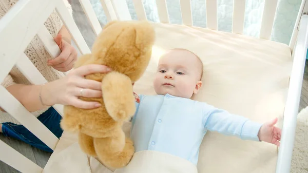 Niño sonriente con los ojos azules sosteniendo y jugando con el oso de peluche en la cuna. Concepto de paternidad, felicidad familiar y desarrollo del bebé — Foto de Stock