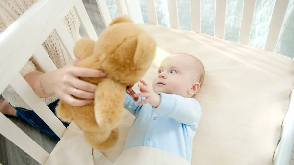 Родитель дарит игрушку медвежонку маленькому мальчику, лежащему в кроватке — стоковое фото