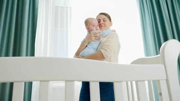 Feliz madre sonriente abrazando y besando a su pequeño hijo por la mañana de pie en el dormitorio con gran ventana. Concepto de paternidad, felicidad familiar y desarrollo del bebé —  Fotos de Stock