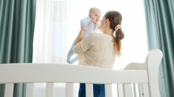 Joven madre de pie en la cuna y sosteniendo a su hijo sonriente. Concepto de paternidad, felicidad familiar y desarrollo del bebé —  Fotos de Stock