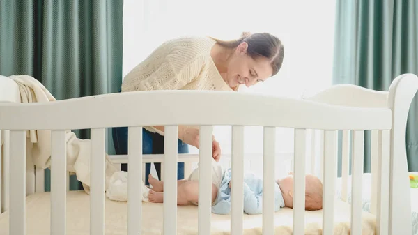 Krásná usměvavá matka svléká svého malého synka ležícího ráno v kolébce, aby vyměnila plenky. Pojem rodičovství, rodinné štěstí a vývoj dítěte. — Stock fotografie