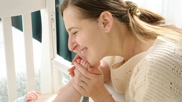 Happy mother looking at her little baby son in bed and kissing tiny feet. Concept of parenting, family happiness and baby development. — Stock Photo, Image