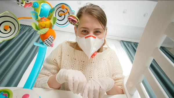 Donna felice indossando maschera medica giocare con il bambino sdraiato nella culla. Concetto di genitorialità e cura del bambino durante la pandemia di coronavirus covid-19. — Foto Stock