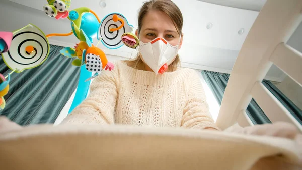 Jeune mère attentionnée en masque médical et gant en caoutchouc couvrant son petit bébé couché dans le berceau. Concept de parentalité et de soins aux bébés pendant la pandémie de coronavirus covid-19. — Photo