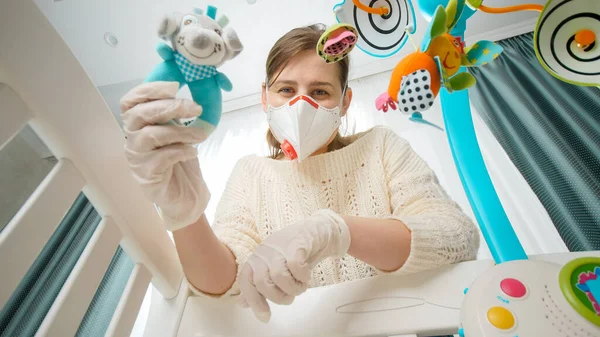 Young mother wearing medical mask and gloves shaking toy rattle with her little baby lying in cradle. Concept of parenting and baby care during coronavirus covid-19 pandemic. — Stock Photo, Image