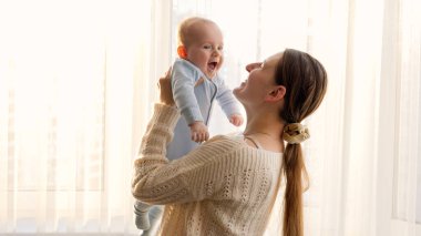Sevimli küçük oğlan, gülümseyen annesine büyük bir pencere ve gün batımı ışığında sarılıyor. Aile mutluluğu ve ebeveynlik kavramı
