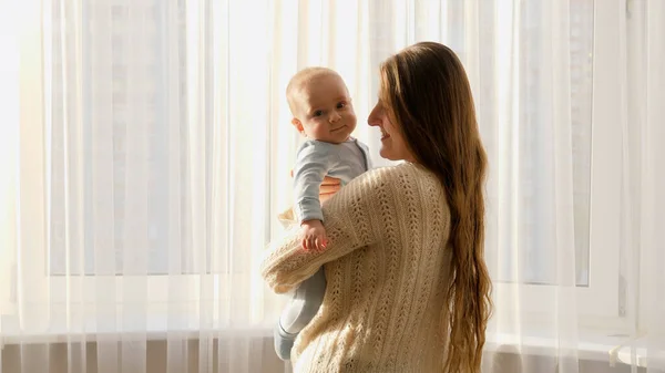 Счастливая улыбающаяся молодая мать с симпатичным маленьким ребенком, стоящим у большого окна с занавесками. Концепция семейного счастья и воспитания — стоковое фото