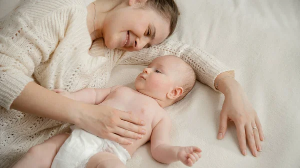 Bella madre sorridente accarezzando delicatamente e guardando il suo bambino accanto a lei a letto. Concetto di genitorialità, cura del bambino e felicità familiare — Foto Stock