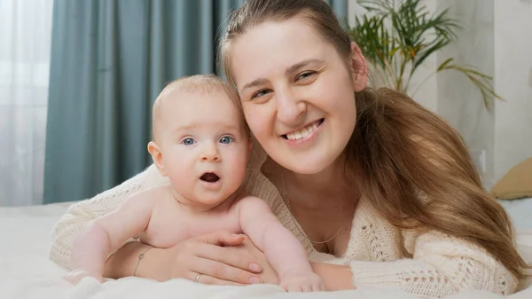 笑顔母抱擁の肖像画彼女の小さな赤ちゃん息子オンベッドと見ますカメラ — ストック写真