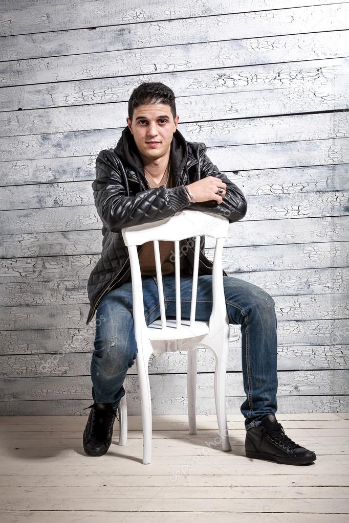 man in jeans and jacket sitting on chair against wooden wall