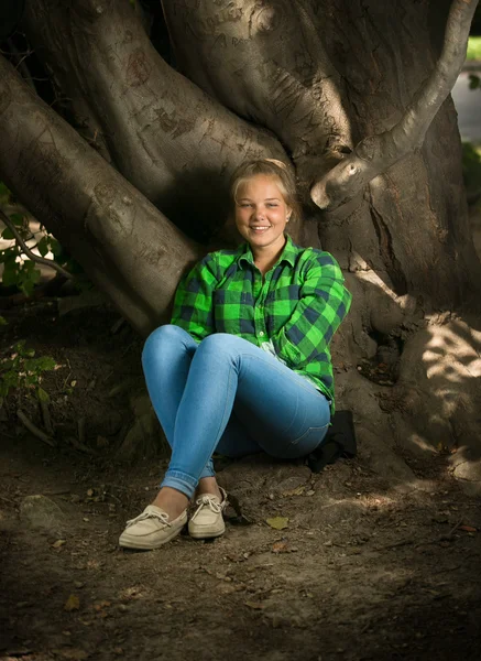 Блондинка в рубашке и джинсах сидит под большим деревом — стоковое фото