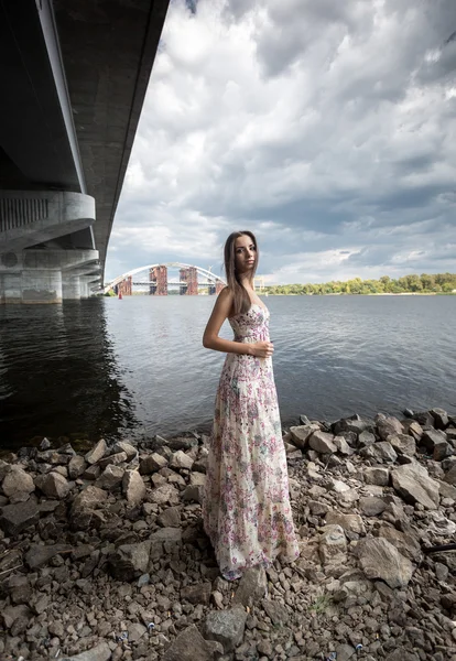 Mujer delgada en vestido posando debajo del puente junto al río — Foto de Stock