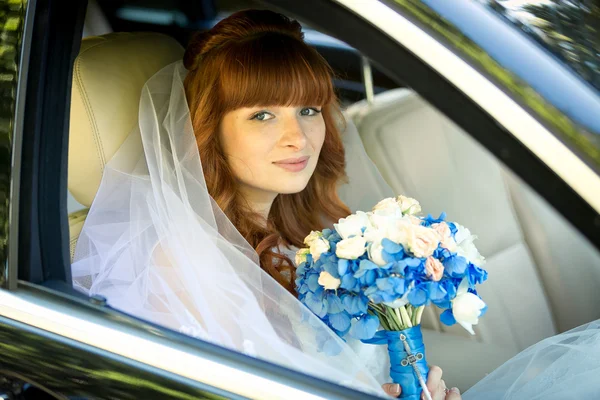 Rödhårig brud sitter i bilen och hålla blå bröllop bukett — Stockfoto