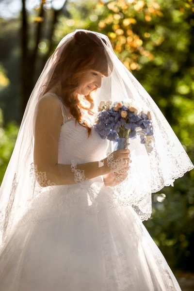 Noiva com véu branco longo olhando para o buquê de casamento — Fotografia de Stock