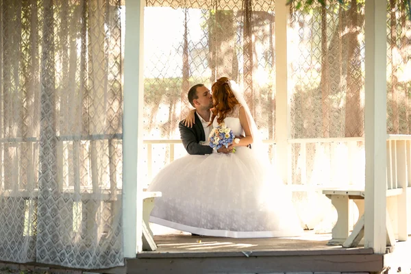 Bride sitting on grooms legs on bench at gazebo — Stock Photo, Image