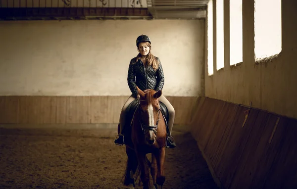 Молодая женщина жокей сидит на коричневой лошади на крытой арене — стоковое фото