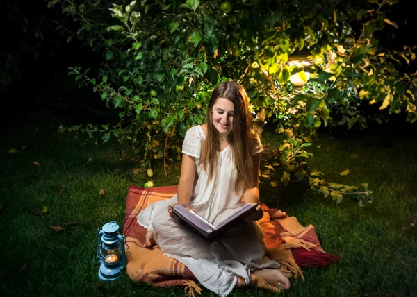 Femme souriante assis la nuit dans le jardin et la lecture grand vieux boo — Photo