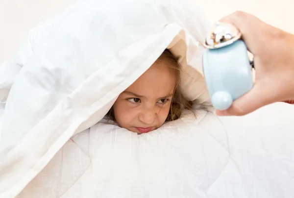 Little girl covering head with pillow and looking at alarm clock — Stock Photo, Image