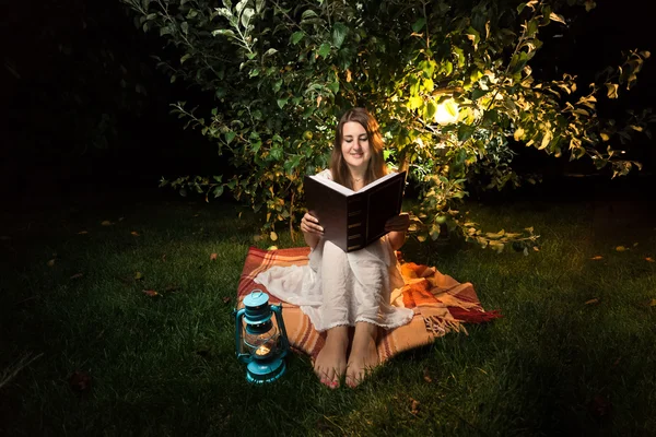 Mujer sonriente sentada en la hierba por la noche y leyendo un gran libro viejo —  Fotos de Stock