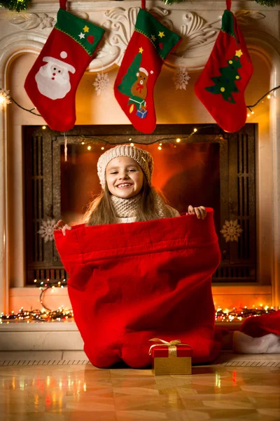 Niña sonriente sentada en bolsa roja para regalos en la chimenea — Foto de Stock