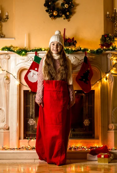 Mädchen steht in santa-roter Tasche im Wohnzimmer neben Kamin — Stockfoto