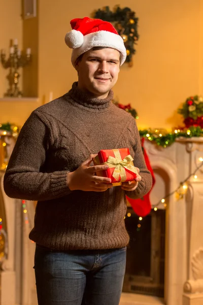 Smiling man in Santa red hat holding red gift box at house — Stock Photo, Image