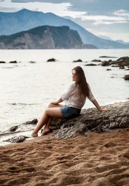 Mujer relajante sentado en la roca grande en la playa y mirando lejos un — Foto de Stock