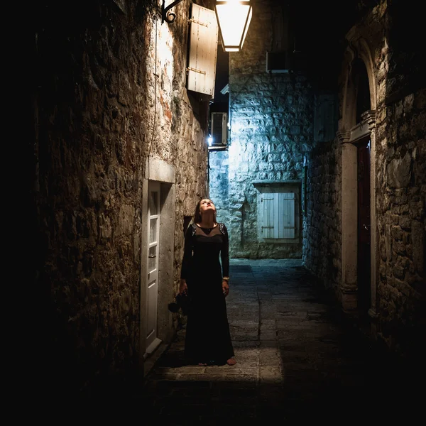 Woman in long black dress standing under lantern at old narrow s — Stock Photo, Image