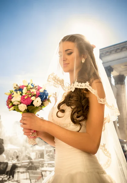 Gelukkig brunette bruid poseren met bruiloft boeket op zonnige dag — Stockfoto