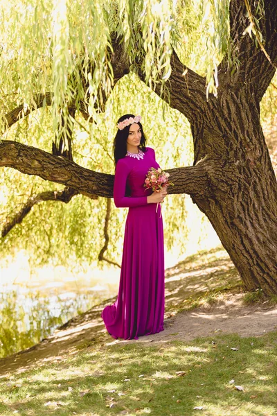 Mujer delgada en vestido púrpura posando en un gran árbol viejo en la orilla del río — Foto de Stock