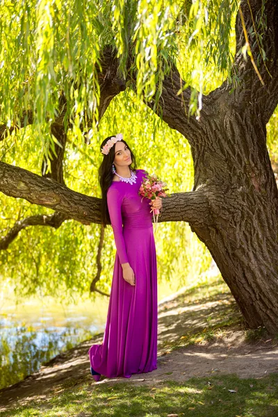 Sexy vrouw in lange jurk leunend tegen een grote boom op de rivieroever — Stockfoto