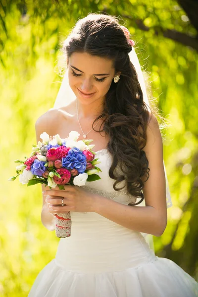 Noiva morena feliz olhando para o buquê de casamento no parque — Fotografia de Stock