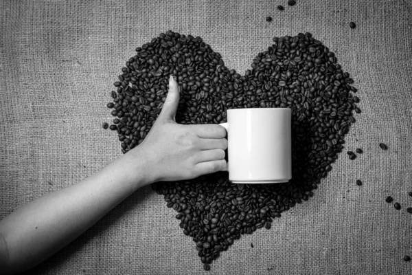 Hand with thumb up holding mug against heart made of coffee bean — Stock Photo, Image