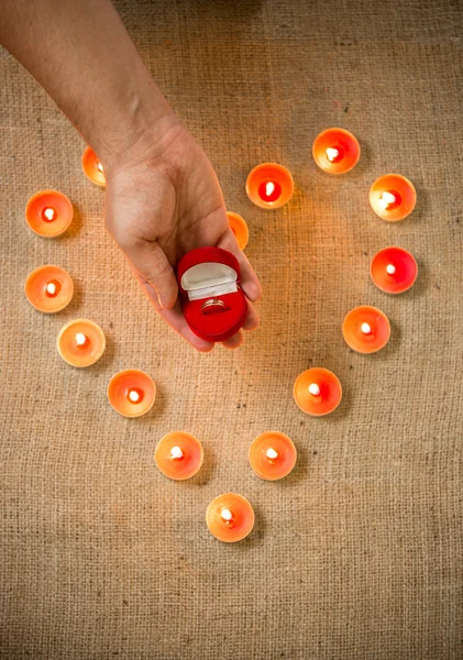 Persona sosteniendo anillo de oro en caja contra corazón hecho de velas — Foto de Stock