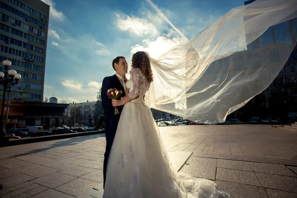 Bride with long veil hugging with groom on street at windy day — Stock Photo, Image