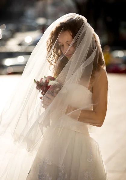 Mignonne mariée bouclée avec long voile regardant bouquet — Photo