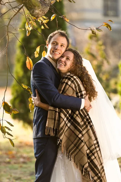 Lachende bruid en bruidegom knuffelen onder plaid in herfst park — Stockfoto