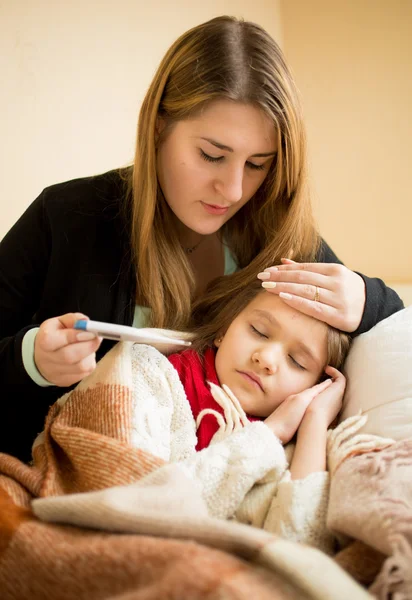 Jovem mãe abraçando filha doente e verificando termômetro — Fotografia de Stock
