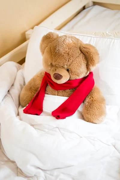 Brown teddy bear in red scarf lying in bed under blanket — Stock Photo, Image