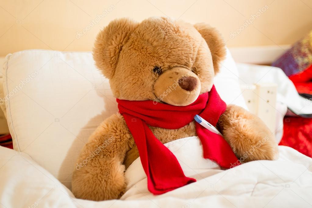 brown teddy bear in read scarf lying in bed with thermometer