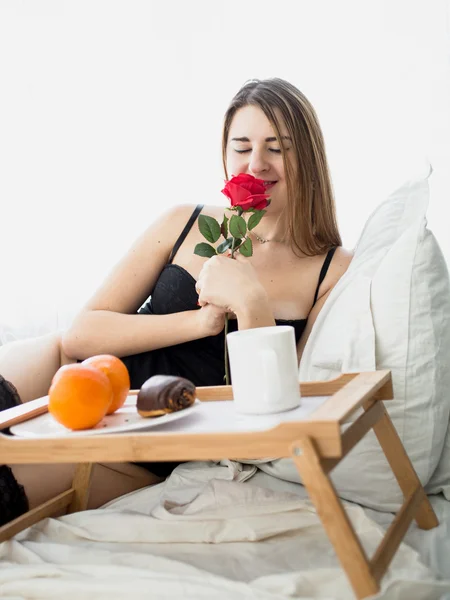 Bela mulher tomando café da manhã na cama e cheirando rosa vermelha — Fotografia de Stock