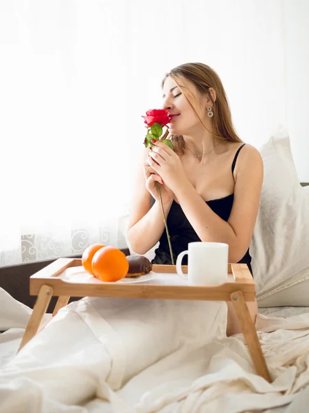 Happy woman smelling rose while having breakfast in bed — Stock Photo, Image