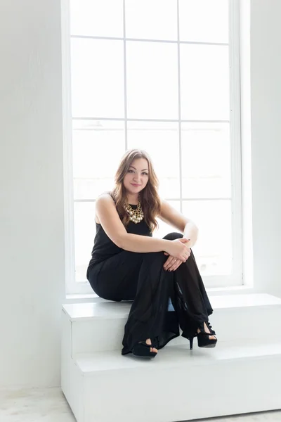 Mujer joven en vestido negro largo sentado en las escaleras en la ventana grande — Foto de Stock