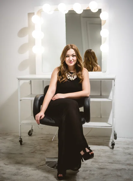 Beautiful woman in long black dress posing in chair against mirr — Stock Photo, Image