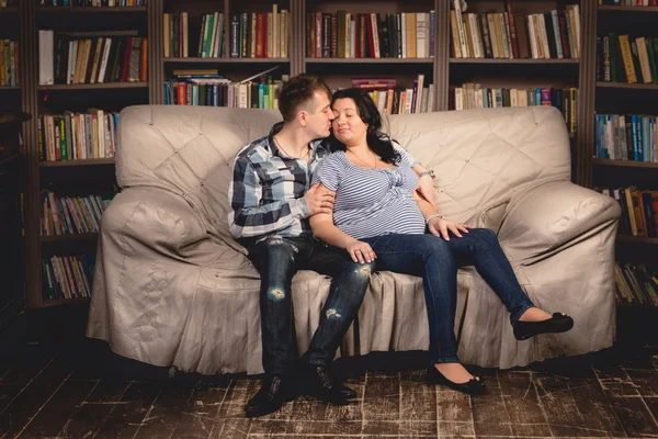 Young pregnant couple hugging on sofa against bookshelves — Stock Photo, Image