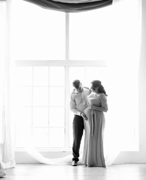 Black and white photo of man hugging pregnant wife against windo — Stock Photo, Image